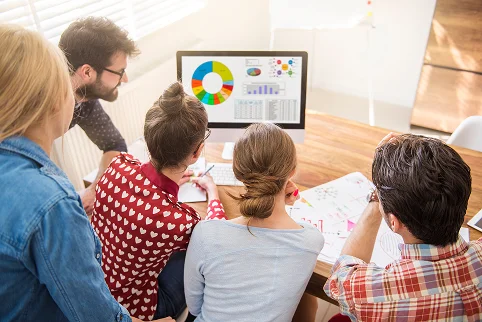 Een groep collega's van achteren gezien die rond een computerscherm zitten en marketinggrafieken analyseren